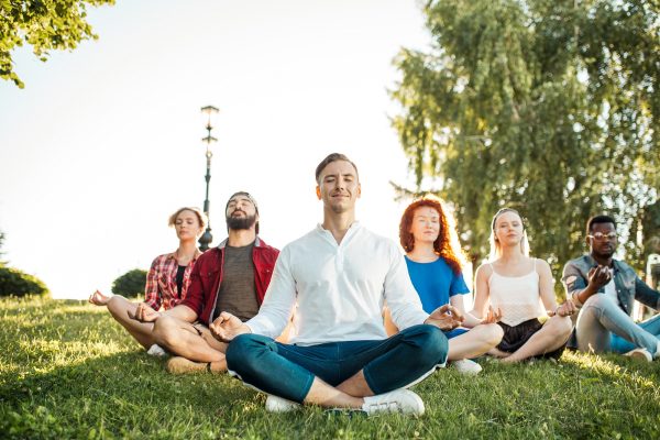 Yoga at park. Interracial friends meditating and spending their leisure time outdoors. Concept of healthy lifestyle, friendship and diversity. Panaromic view of meditating group over nature background
